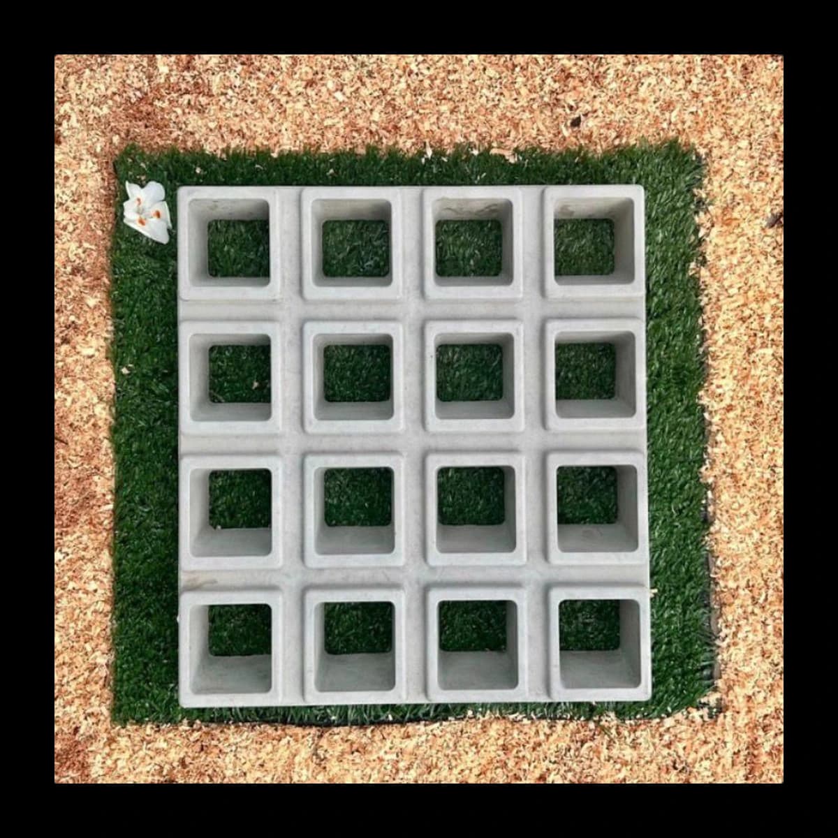 Cobogó modelo 16 Furos 3D em concreto pré-moldado · módulo individual com 16 furos quadrados em arranjo 4 por 4, paredes em profundidade tipo caixa criando efeito tridimensional, vista isolada sobre tapete de grama sintética com flor branca decorativa · Central do Concreto Ivoti RS