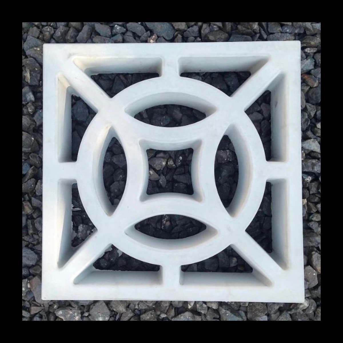 Cobogó modelo Eclipse em concreto pré-moldado · módulo individual quadrado com círculo central inscrito e quatro pétalas curvas vazadas formando padrão simétrico tipo bússola, vista isolada sobre fundo de brita escura · Central do Concreto Ivoti RS