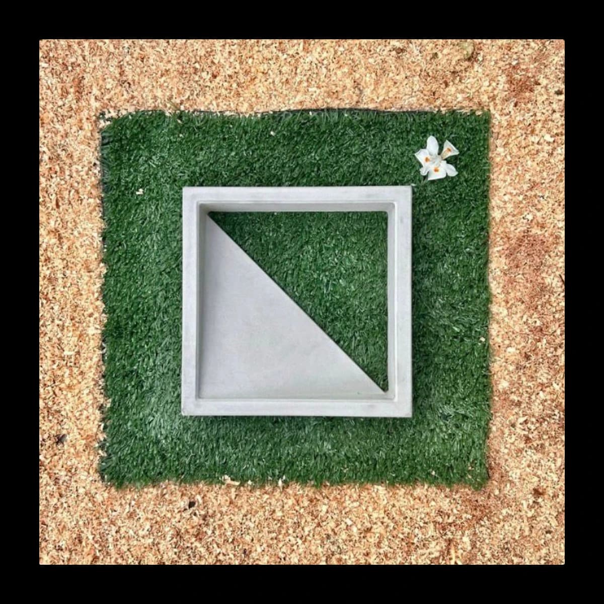 Cobogó modelo Urbano em concreto pré-moldado · módulo individual quadrado com diagonal triangular vazada, vista isolada sobre tapete de grama sintética com flor branca decorativa · Central do Concreto Ivoti RS