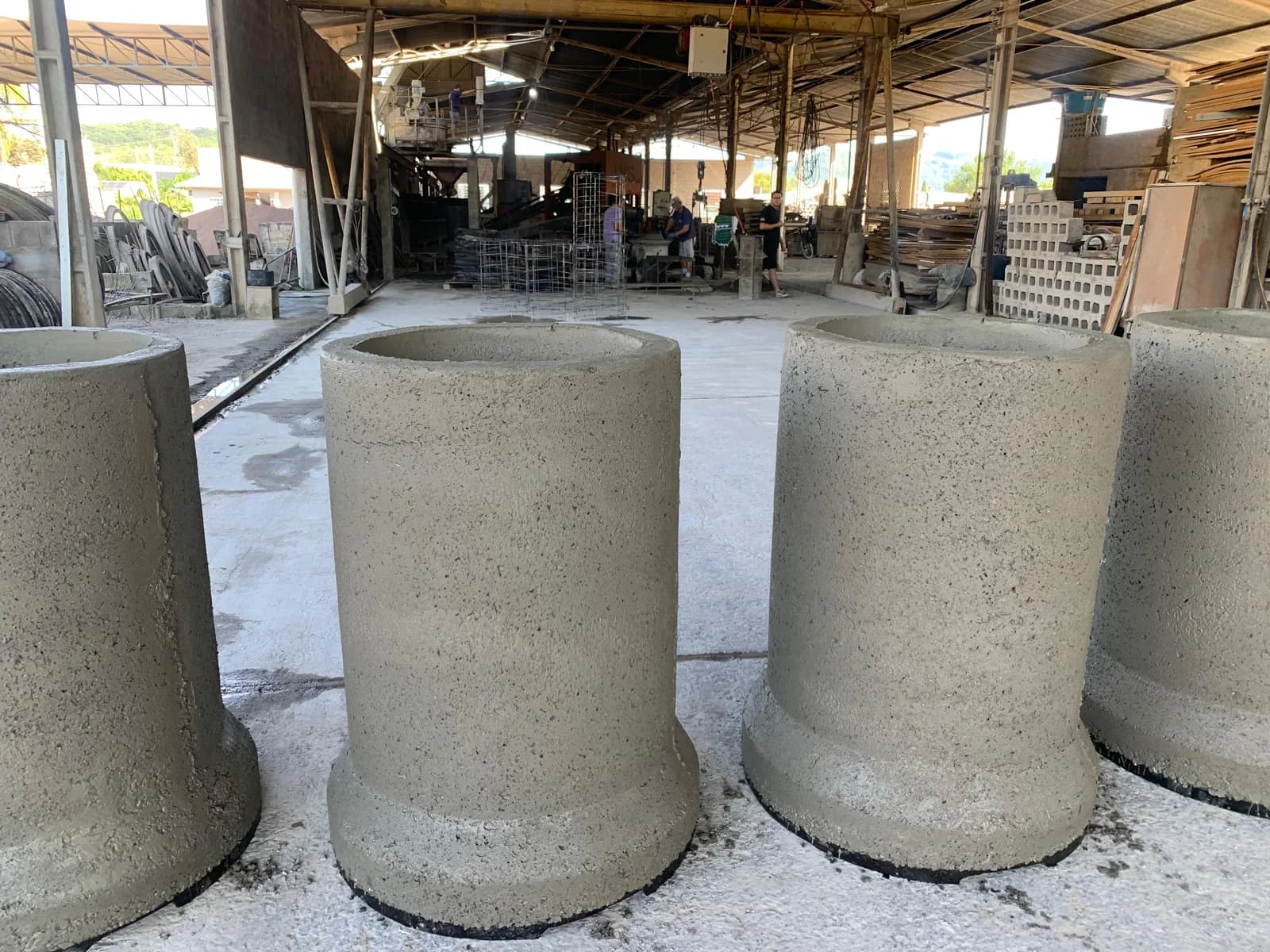 Tubos de concreto em pé no piso de um galpão de produção de pré-moldados, com gaiolas de armadura e blocos de concreto ao fundo