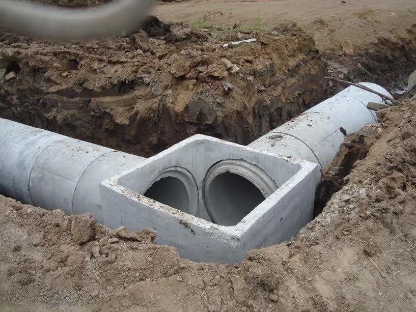 Caixa de passagem de concreto conectada a dois tubos instalados em vala de drenagem pluvial em obra real — aplicação típica do tubo de concreto.